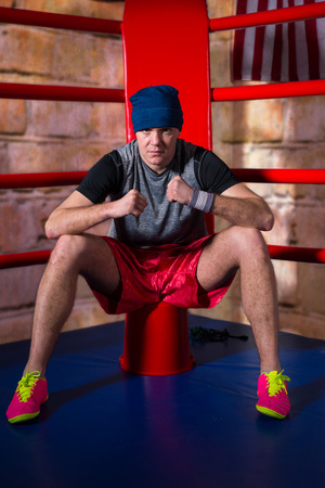 Athletic young male boxer sitting near red corner of a regular boxing ring surrounded by ropes in a gymの写真素材