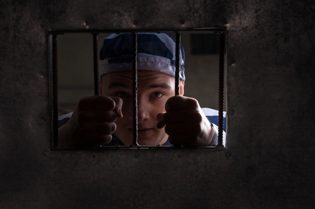 View through iron door with prison bars on male prisoner holding bars wearing prison uniform in a jail cellの写真素材