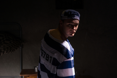Male prisoner wearing prison uniform looked back standing near his bed in a small dark prison cellの写真素材