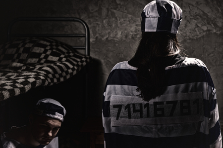 Female prisoner wearing prison uniform with sewed number standing with her back near male prisoner sitting on a bed in a jail cellの写真素材