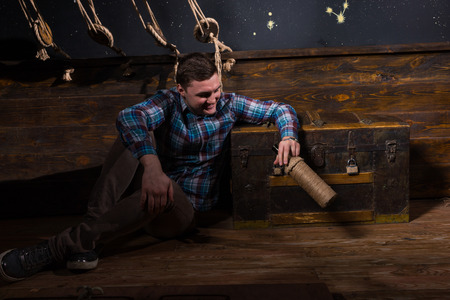 A young male sits near a chest, looking into glass bottle and trying to get out of the trap, escape the room game conceptの写真素材