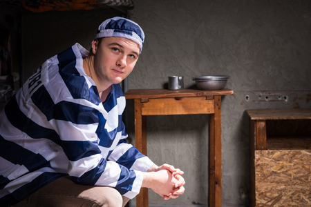 Young man wearing prison uniform sitting on a bed near bedside table with aluminum dishes in a small prison cellの写真素材