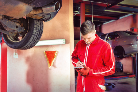 Young male motor mechanic standing making notes underneath a lifted car near working equipment for...