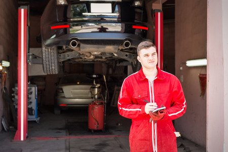Thoughtful motor mechanic standing making notes in front of a black sedan elevated on a hoist in a bay in a garage or workshopの写真素材