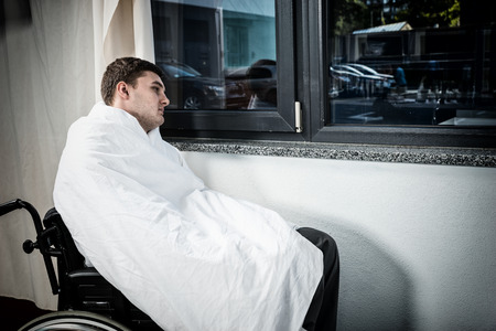 Sad male ill patient sitting by the window on wheelchair covered with quilt in hospital ward. Healthcare conceptの写真素材