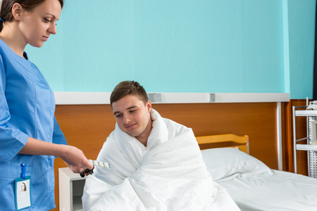 Young attractive nurse giving pills to ill patient sitting on wheelchair covered with quilt in hospital ward. Healthcare conceptの写真素材