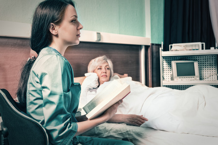 Attractive nurse in uniform is reading a book for her patient who is lying in the hospital bed in the hospital ward. Healthcare conceptの写真素材