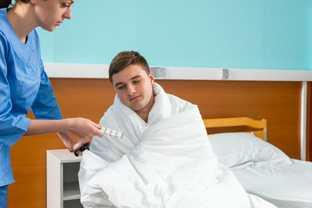 Young nurse giving pills to ill patient sitting on wheelchair covered with quilt in hospital ward. Healthcare conceptの写真素材