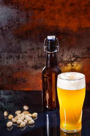 Glass with freshly poured unfiltered light beer, beer bottle near scattered pistachios on a black mirror surface. Food and beverages conceptの写真素材