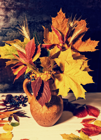 Beautiful clay jug with autumn leaves and wheat near scattered leaves on white wooden desk. Thanksgiving day and autumn conceptの写真素材