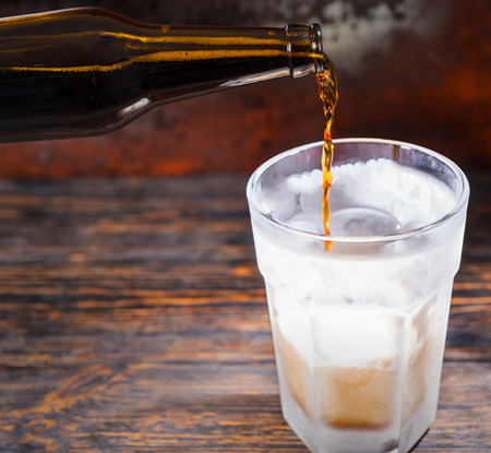 Beer is pouring into a frozen glass from brown bottle on dark wooden desk. Food and beverages conceptの写真素材