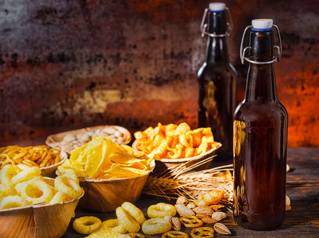 Wooden plates with snacks near two bottles of beer, wheat, scattered nuts and pretzels on dark wooden desk. Food and beverages conceptの写真素材
