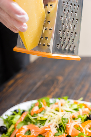 Hands in gloves rub cheese on a grater above salad with mix of lettuce leaves, shrimps, salmon and olives in white plate on wooden deskの写真素材