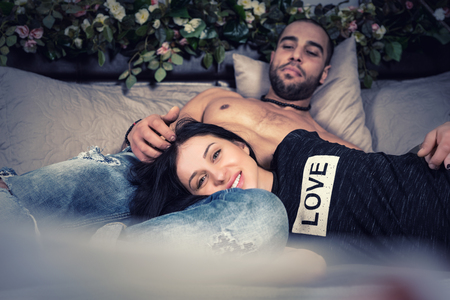 Wonderful happy couple of man with bare chest and brunette pregnant woman lying on the gray cozy bed with flowers on the bedstead in the bedroomの写真素材