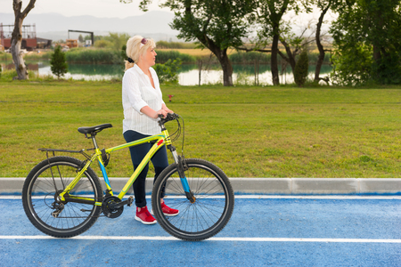 Senior woman stood with bicycle on track with grass and lake in background.の写真素材