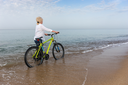 Blond woman in jeans walking in the sea wheeling a bicycle along a sandy shoreline on a misty day at the beach with copy spaceの写真素材