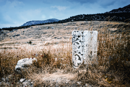 Remnants of a carved classical column in long grass in a field in a mountainous landscape from an ancient historical ruinの写真素材