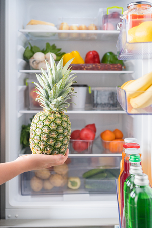 Person holding fresh pineapple against open fridge full of foodの写真素材