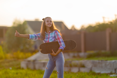 Cute young teenage girl hailing a lift standing at the edge of a road in the warm glow of the sunset thumbing a ride with a skateboard under her armの写真素材