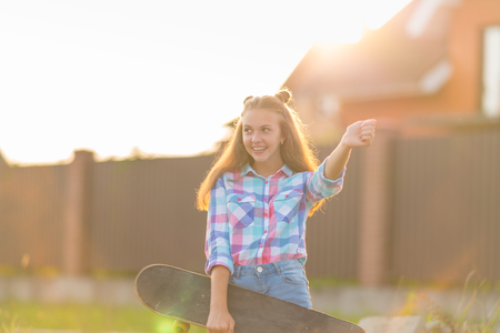 Cute happy young woman with a skateboard under her arm standing waving with a smile in the glow of the sunsetの写真素材