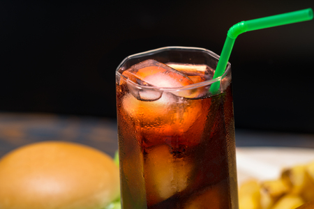 Close up on soda glass with ice small ice cubes and green straw stuck in it while burger and fries sit in backgroundの写真素材