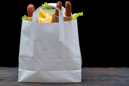 White bag filled with hot dogs and lettuce leaves sitting on wooden table in front of black backgroundの写真素材
