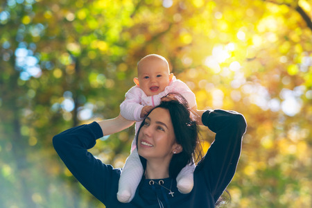Vivacious laughing little baby girl enjoying a piggy back ride on the shoulders of her mother in an autumn forestの写真素材