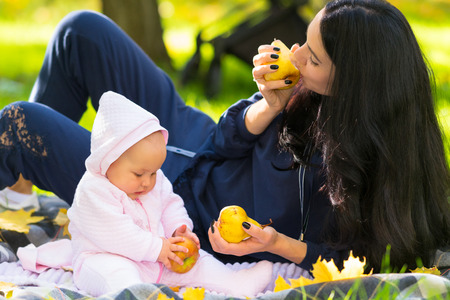 Young mother eating autumn apples with her baby daughter as they relax together on a blanket in a...