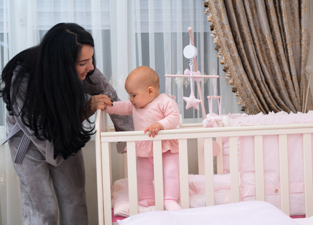 Loving young mother teaching her adorable little baby daughter to stand helping her balance in a corner of her cot in the nurseryの写真素材