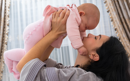 Loving mother playing with her little baby girl lifting her on high in a close up tender indoor...