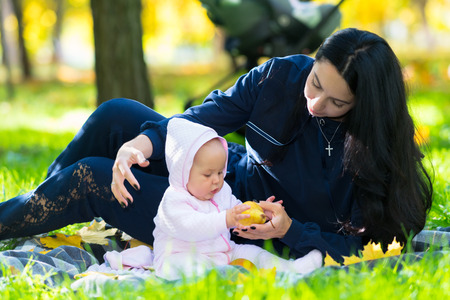 Baby girl playing with an autumn apple as she relaxes outdoors in a park on a rug alongside her...