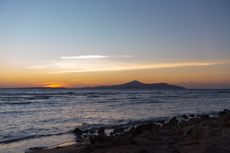 Orange sunset over a tropical island viewed across a calm blue ocean from a rocky shoreline and beachの写真素材