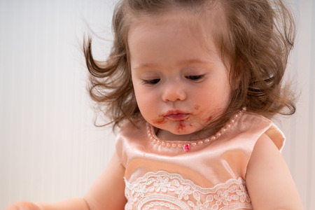 Cute little girl with a dirty sticky face full of red jelly from eating her birthday cake in a close up cropped portraitの写真素材