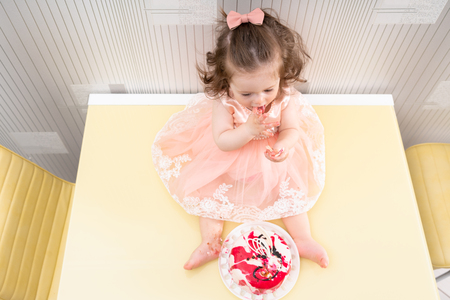 Little baby girl in pink dress sitting in front of the cake on yellow table and eating it with her hands, viewed from aboveの写真素材