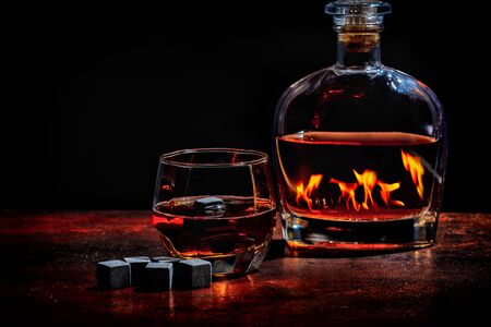 Bottle of cognac or whiskey reflecting a the fames of a hot fire in winter with a glass of sprits and black reusable ice cubes or chilling cubesの写真素材