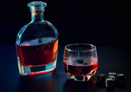 Bottle of cognac or whiskey with full glass and black chilling cubes or reusable ice cubes on a dark...