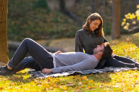 Young woman relaxing with her husband lying on a rug on the grass in evening light in a park in...