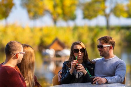 Two couples enjoying coffee at an outdoor restaurant seated at a table overlooking tranquil water in...