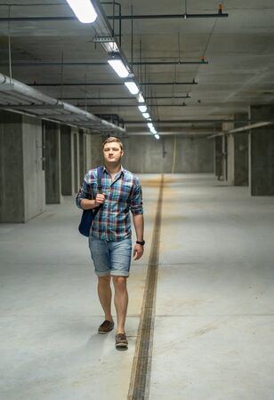 Fit young man walking through a bare empty concrete basement along the metal drain approaching the camera carrying a backpack over his shoulderの写真素材