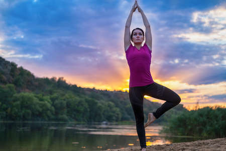 Young woman doing the yoga tree pose at sunset backlit by a colorful orange sky reflecting on tranquil water in nature in a health and fitness or active lifestyle conceptの写真素材