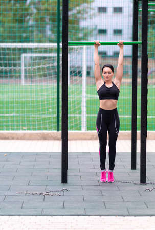 Fit sporty young woman exercising on a parallel bar at an outdoor gym and sports field hanging from...