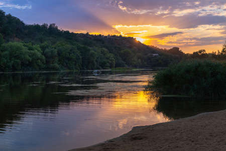 Tranquil fiery orange sunset with reflections on calm water of a lake or river surrounded by trees viewed from a sandy beachの写真素材