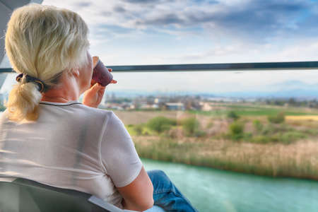 Woman sitting enjoying a drink on an outdoor patio facing out through a glass balustrade overlooking...
