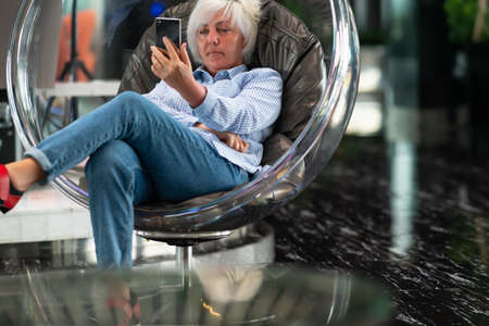 Woman engrossed in watching media on her smartphone relaxing in a modern transparent perspex tub...