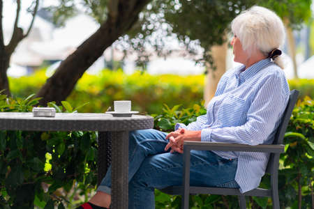 Pensive woman relaxing on an outdoor patio at a restaurant table with a cup of tea as she stares out over the lush leafy green plants at the scene outsideの写真素材