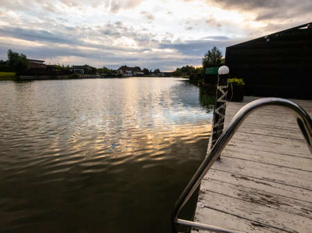 View over a jetty of a tranquil lake at sunset with houses lining the shoreline silhouetted against the skyの写真素材
