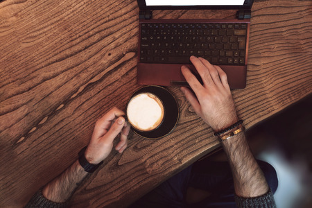 Hands of a young man, a cup of coffee and a laptop at a wooden table in a cafeの写真素材