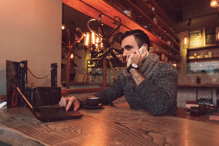 A young man with a beard sits at a table in a cafe and works behind a small laptop with a cup of...