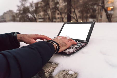 A young guy with a beard working on a laptop in the winter on the street is very cold computer all...