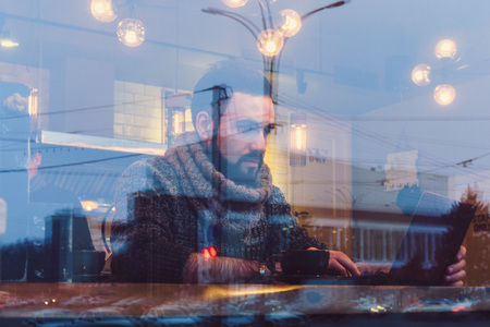 A young guy with a beard in a cozy cafe sits behind a panoramic glass with a laptop and is engaged...